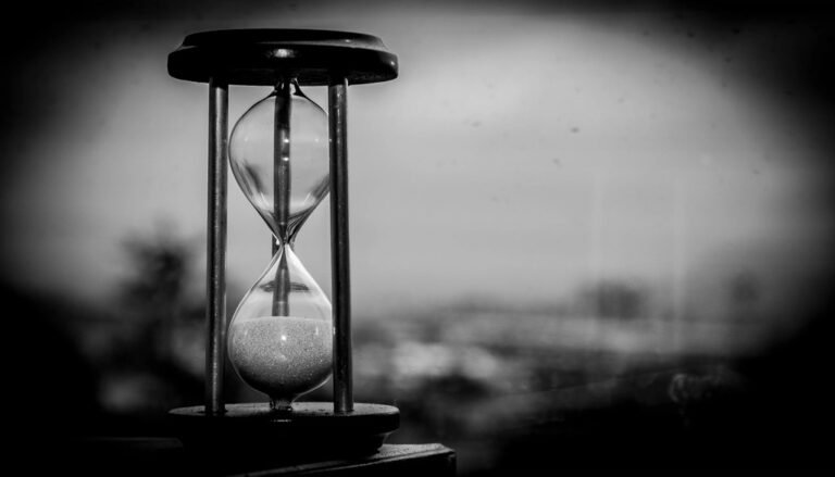 A black and white hourglass with most of its sand already run through, against a blurred window