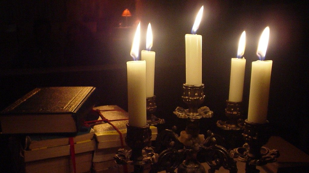 Lit candles on a candelabra beside stacks of old books in warm light against darkness