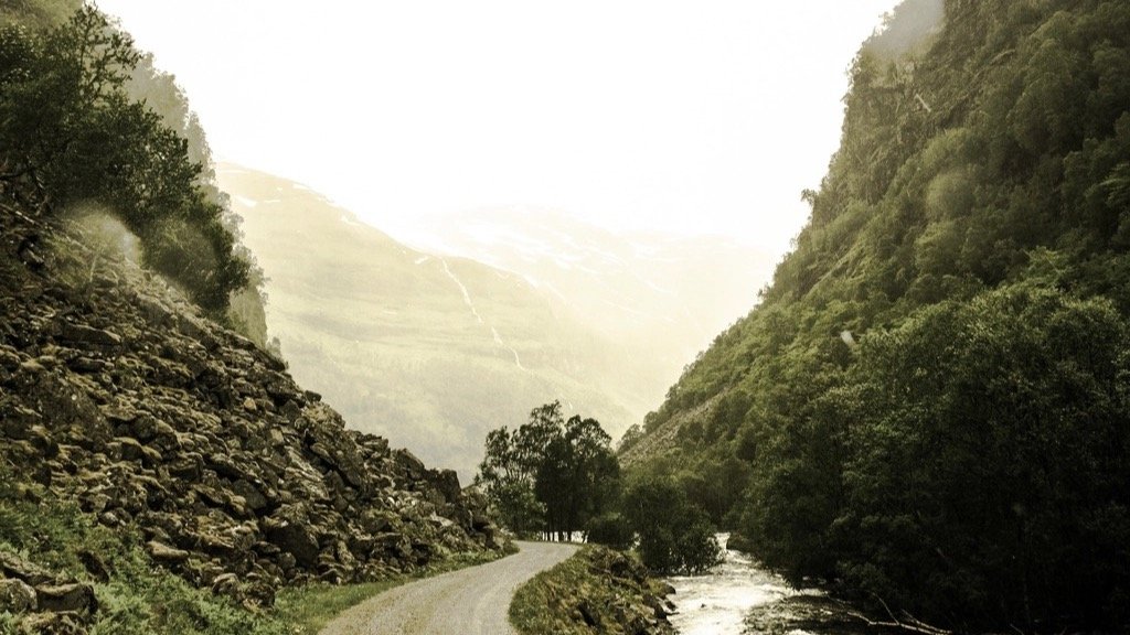 A gravel road winding through a green mountain valley toward bright light