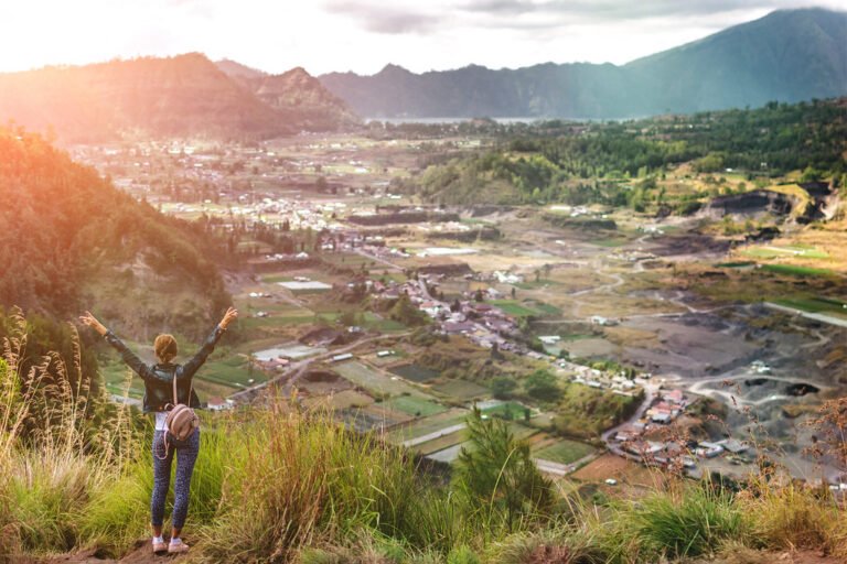 A woman standing on a mountaintop with arms raised, overlooking a sunlit valley